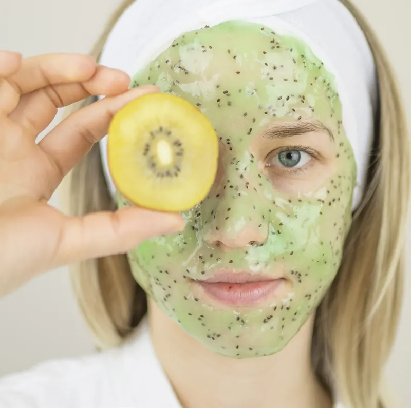 Femeie cu mască verde esthemax® cu kiwi, ținând o jumătate de kiwi